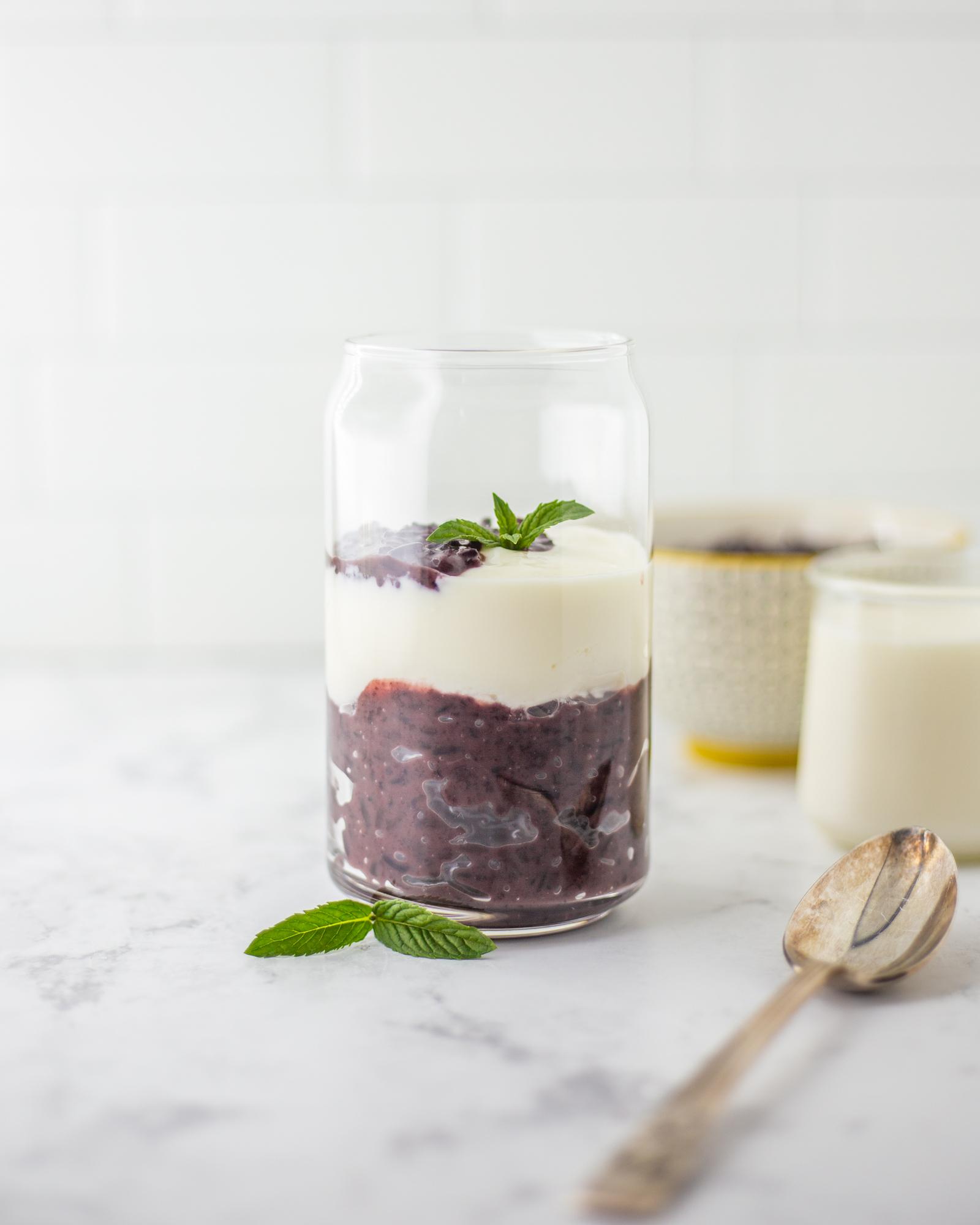 A glass filled with layers of creamy yogurt and purple sticky rice, garnished with mint leaves.